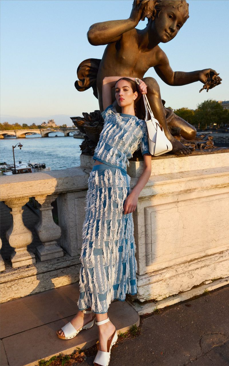 Fashion shot of a woman in a blue and white outfit posing on a bridge near a bronze statue, with a river and buildings in the background.