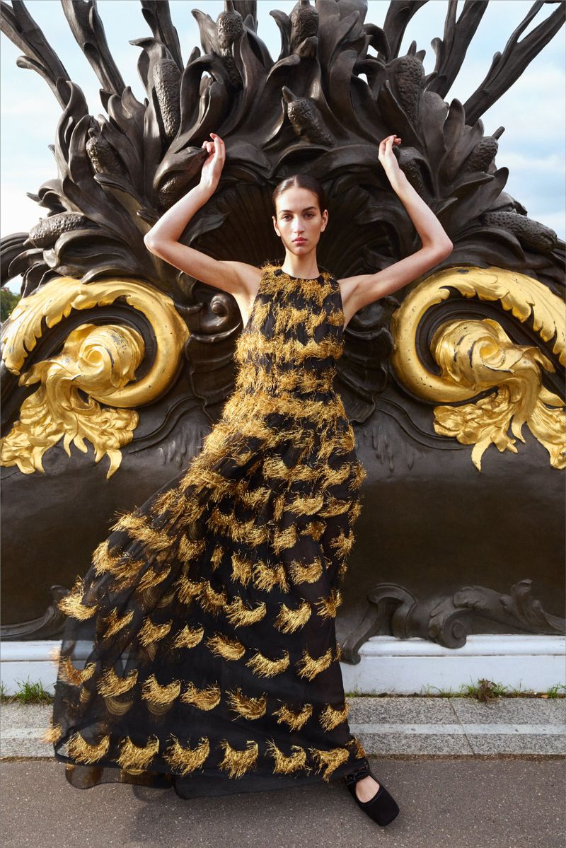 Woman in black and gold gown posing outdoors with arms raised near a decorative fountain with an overcast sky in the background.