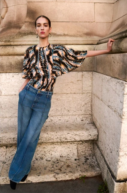 Adult female fashion model in a patterned top and denim jeans poses against a light stone wall in natural outdoor lighting.
