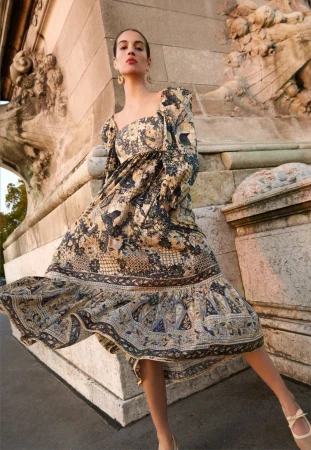 Elegant woman in a patterned midi dress poses by a stone archway in golden hour light, showcasing fashion in an outdoor setting.