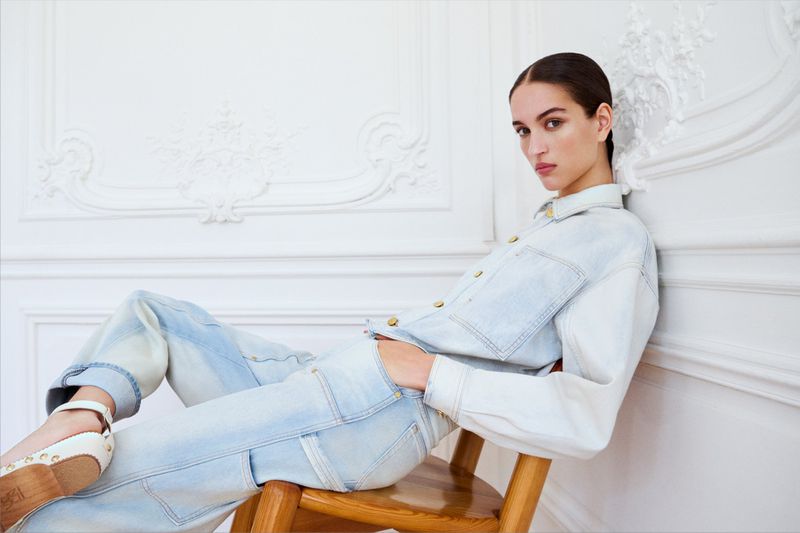 Fashionable woman in a light blue denim outfit, stylish shoes, sitting on a wooden chair against a white wall with ornamental molding.