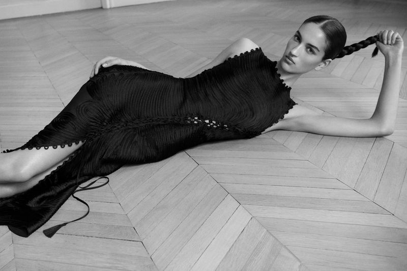 Monochrome fashion shot of a woman in a textured black dress lying on a light wood herringbone floor with a long braid.