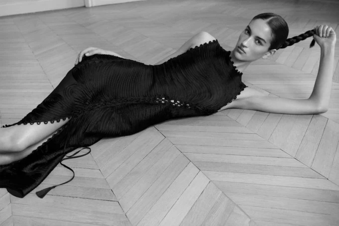 Monochrome fashion shot of a woman in a textured black dress lying on a light wood herringbone floor with a long braid.