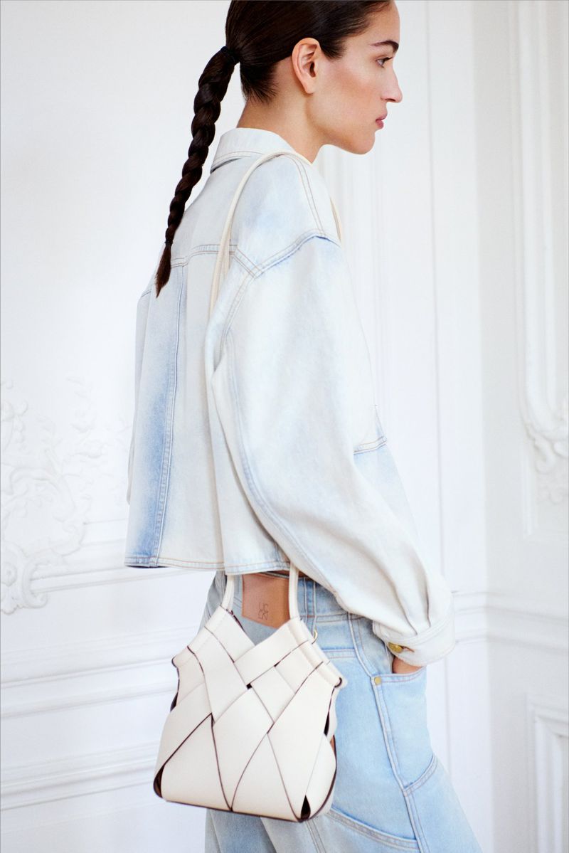 Model Lily Vander Meeden seen from behind in a light-wash denim jacket, holding a white woven leather handbag against a white wall.