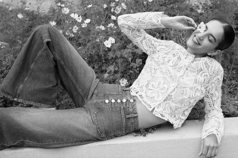 A fair-skinned woman with dark hair lies on a stone surface near white flowers, wearing wide-leg jeans and a lace blouse in a black-and-white image.