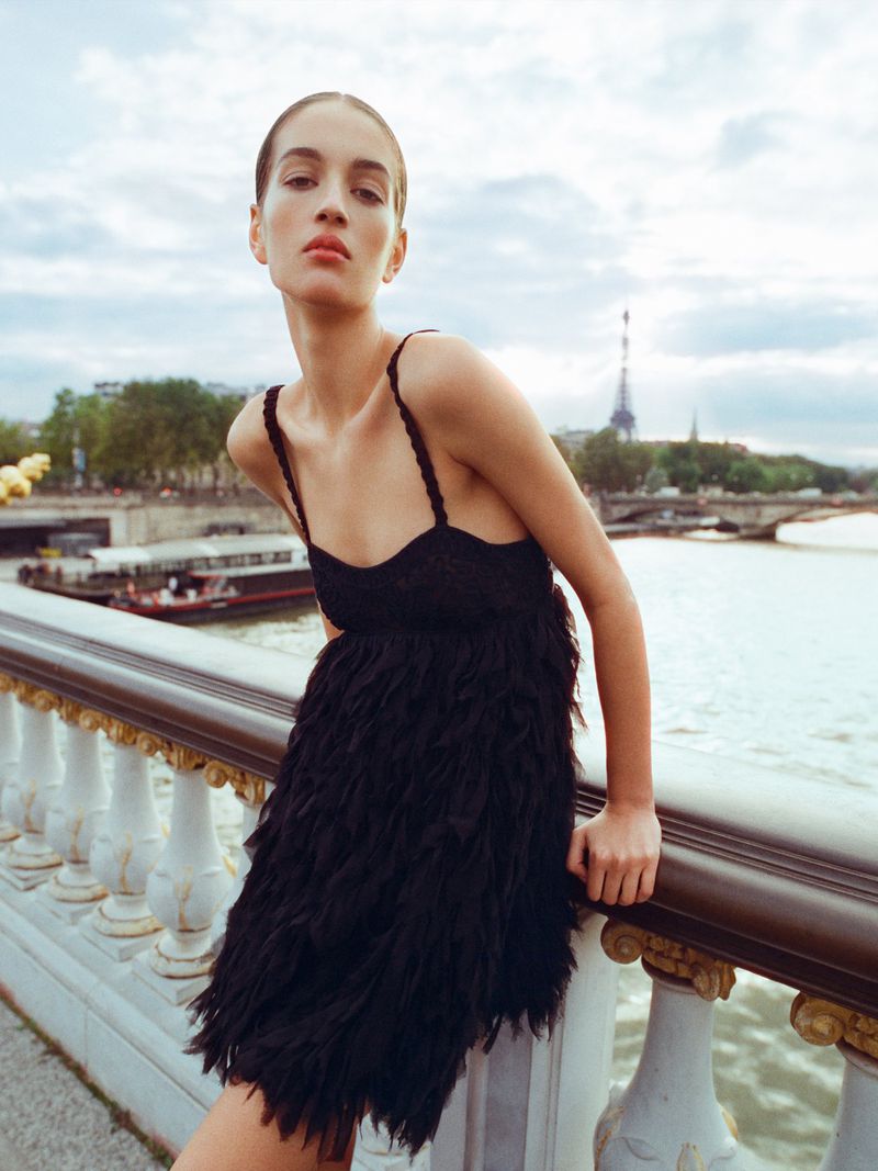 Woman in a black dress standing on a bridge in Paris, France, with the Eiffel Tower in the background on a cloudy day.