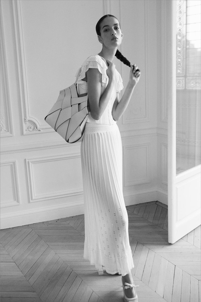 Monochrome portrait of a slender woman in a white dress, holding her braid and carrying a woven tote bag, posing in a bright indoor space.