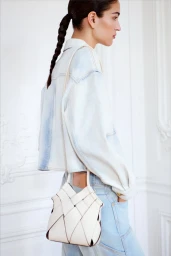 Model Lily Vander Meeden seen from behind in a light-wash denim jacket, holding a white woven leather handbag against a white wall.