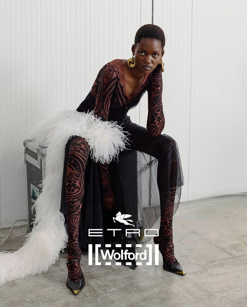 Fashion shot of Black woman seated with Etro and Wolford logo. She wears a printed top, sheer pants, gold earrings, and feather boa.