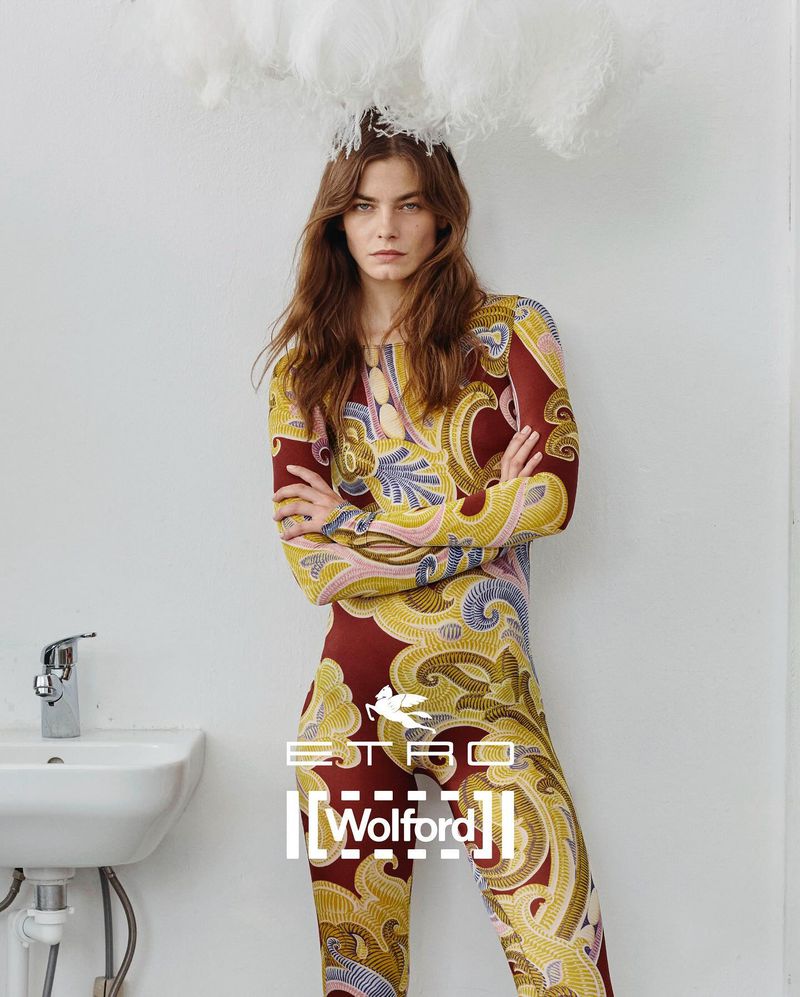 Fashion model in a patterned jumpsuit stands indoors with a cloud-like prop above her head, and sink in the background.