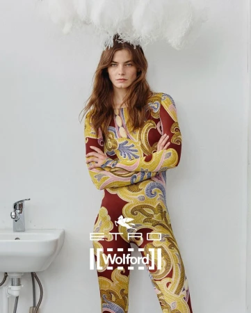 Fashion model in a patterned jumpsuit stands indoors with a cloud-like prop above her head, and sink in the background.