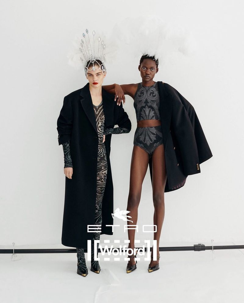 Two fashion models pose in a studio. One wears a long coat and patterned dress with a feathered headpiece; the other wears a two-piece set.