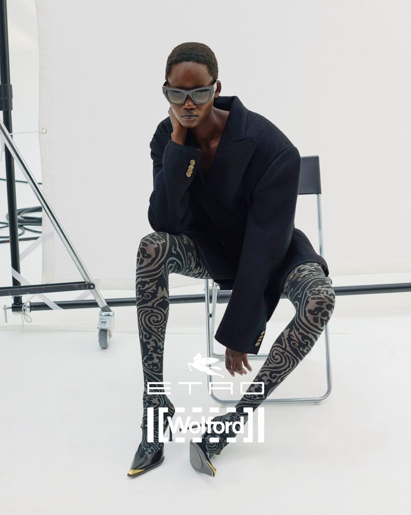 Editorial fashion shot of a woman wearing a black blazer, patterned tights, and black high heels while seated in a photo studio.