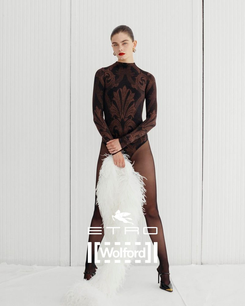 Fashion shot of a woman in patterned bodysuit and sheer tights, holding a feathered stole, posing against a white wall.
