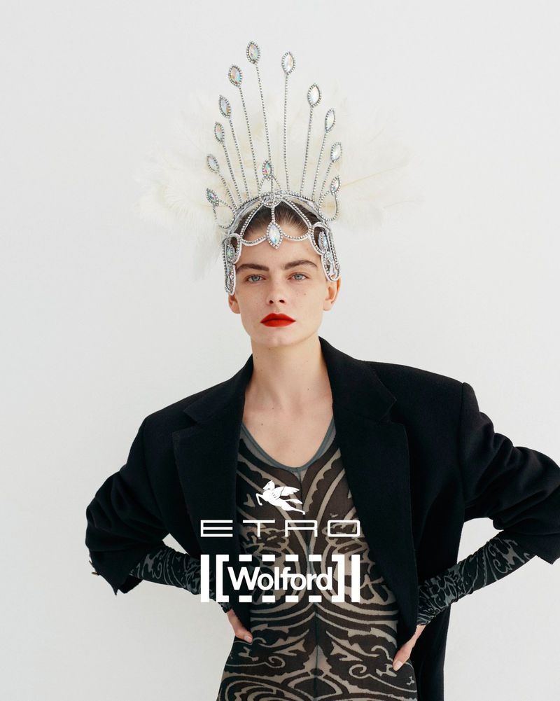 Close-up of a fair-skinned fashion model wearing an ornate jeweled headpiece, dark blazer, and patterned top against a white background.