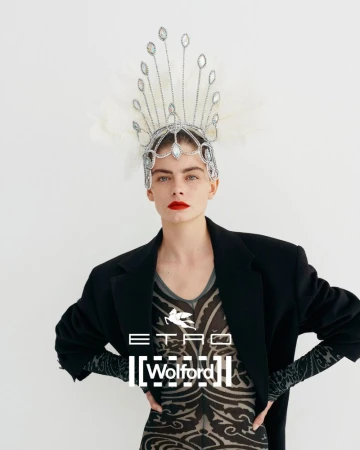 Close-up of a fair-skinned fashion model wearing an ornate jeweled headpiece, dark blazer, and patterned top against a white background.