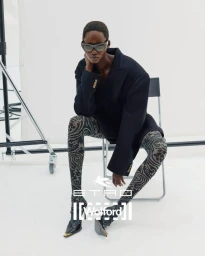 Model wearing patterned tights and a dark blazer seated on a folding chair in front of a white studio backdrop.