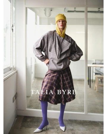 A young, fair-skinned woman poses in a gray plaid top and plaid skirt, accessorized with a yellow hat and scarf, plus purple tights and white heels.