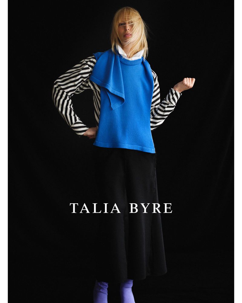Fashion model poses in a studio wearing a bright blue sweater, a black skirt, and a striped long-sleeve top against a dark backdrop.