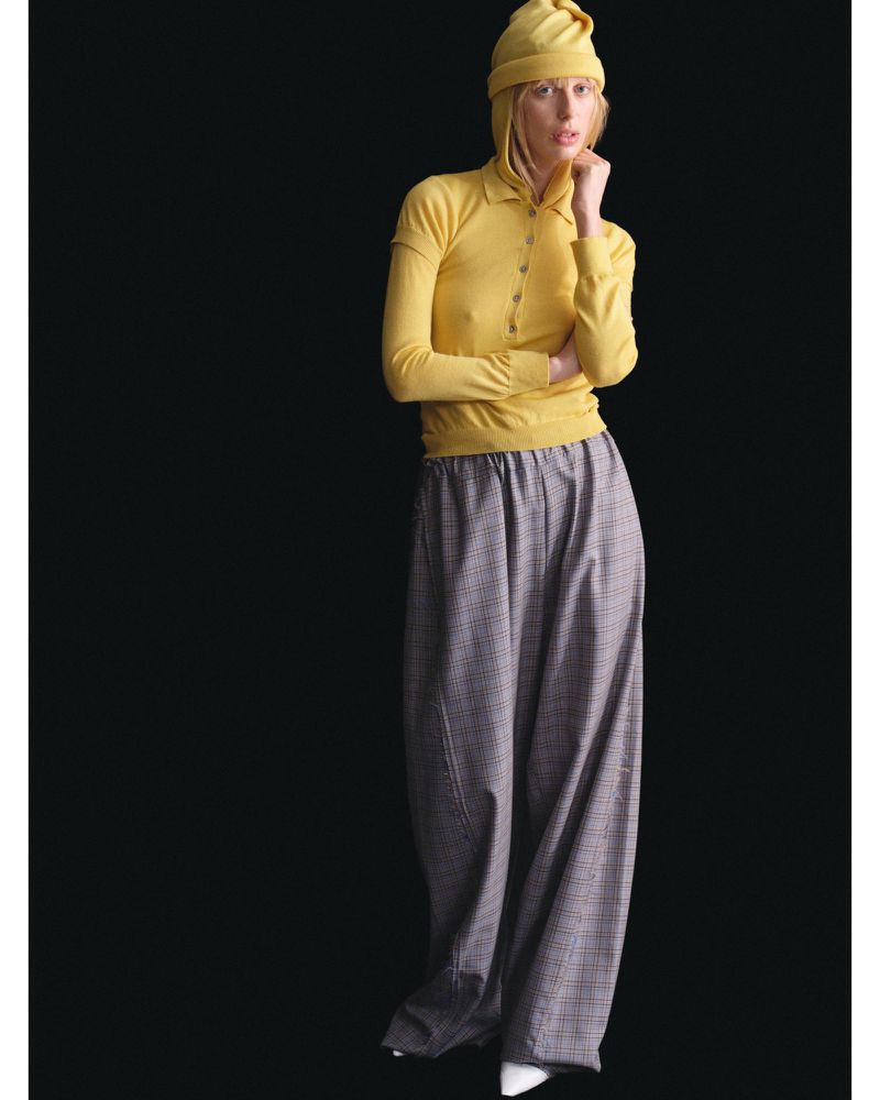 A female fashion model wearing a yellow long-sleeved polo shirt and yellow beanie, paired with oversized plaid pants, stands in a studio.