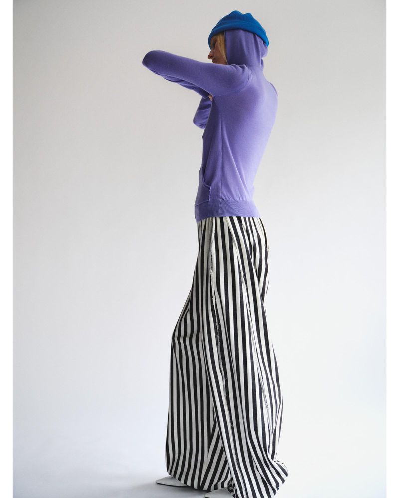 Model Lili Sumner wears a purple hooded top, blue beanie, and black-and-white vertically striped wide-leg trousers against a white backdrop.