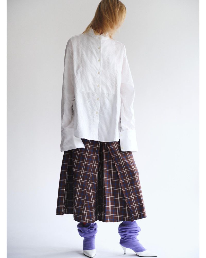 Woman wearing a white shirt, brown plaid skirt, and purple leg warmers against a white backdrop in a fashion editorial shot.