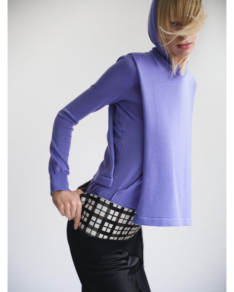 Fashionable adult woman poses in a periwinkle hooded sweater and black skirt against a neutral backdrop in a studio setting.