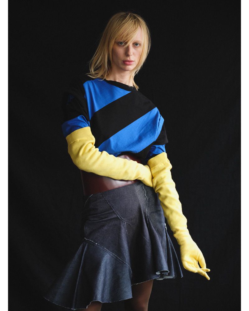 Lili Sumner wearing a blue and black striped top, yellow knit gloves, and a flared denim skirt against a black studio background.
