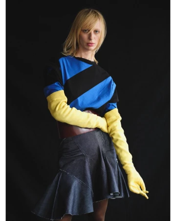 Lili Sumner wearing a blue and black striped top, yellow knit gloves, and a flared denim skirt against a black studio background.