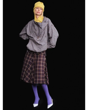 Woman in a grey plaid jacket, brown skirt, and yellow hat posing in front of a black backdrop in a fashion editorial style.