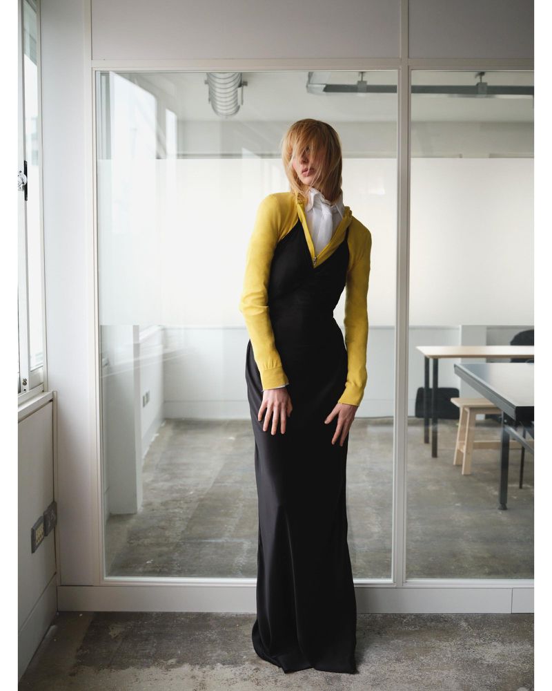 Model Lili Sumner wearing a long black slip dress over a yellow knit top and white collared shirt in front of glass office partitions.