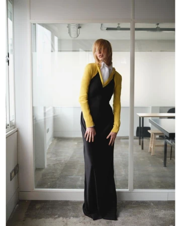 Model Lili Sumner wearing a long black slip dress over a yellow knit top and white collared shirt in front of glass office partitions.