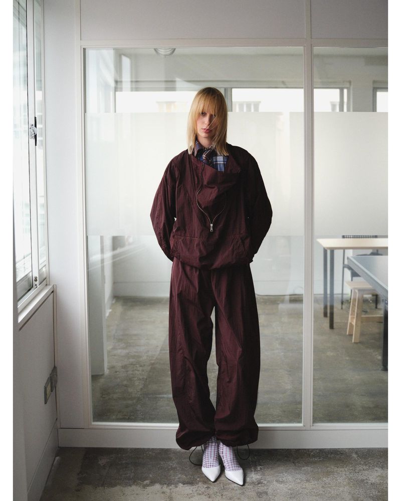 Woman in a maroon tracksuit and white heels stands in front of a glass wall, creating a fashion editorial in an indoor setting.