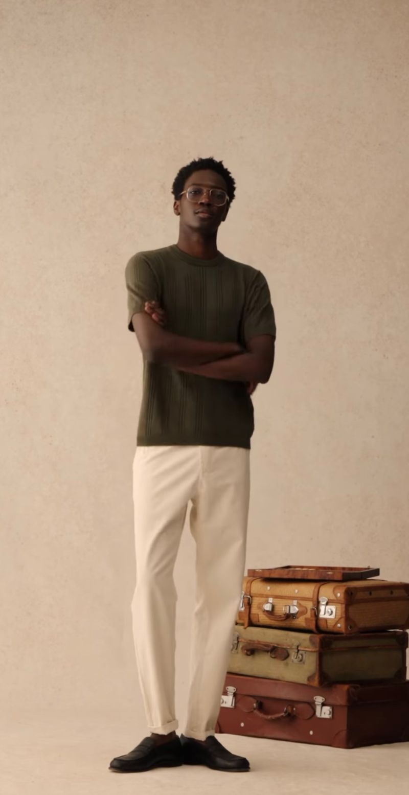 Full shot of a Black male model in green short-sleeve knit shirt and white pants stands next to stacked vintage suitcases on beige background.