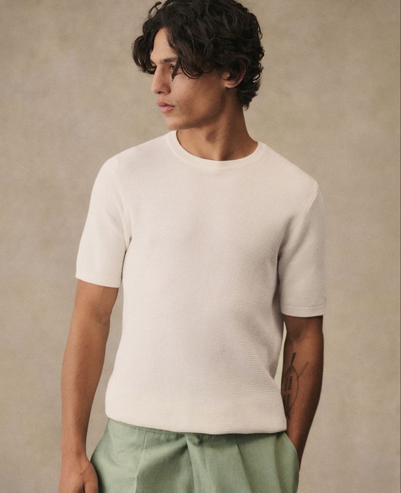 Man wearing a textured white short-sleeve shirt and light green linen pants in a neutral indoor setting, fashion portrait.
