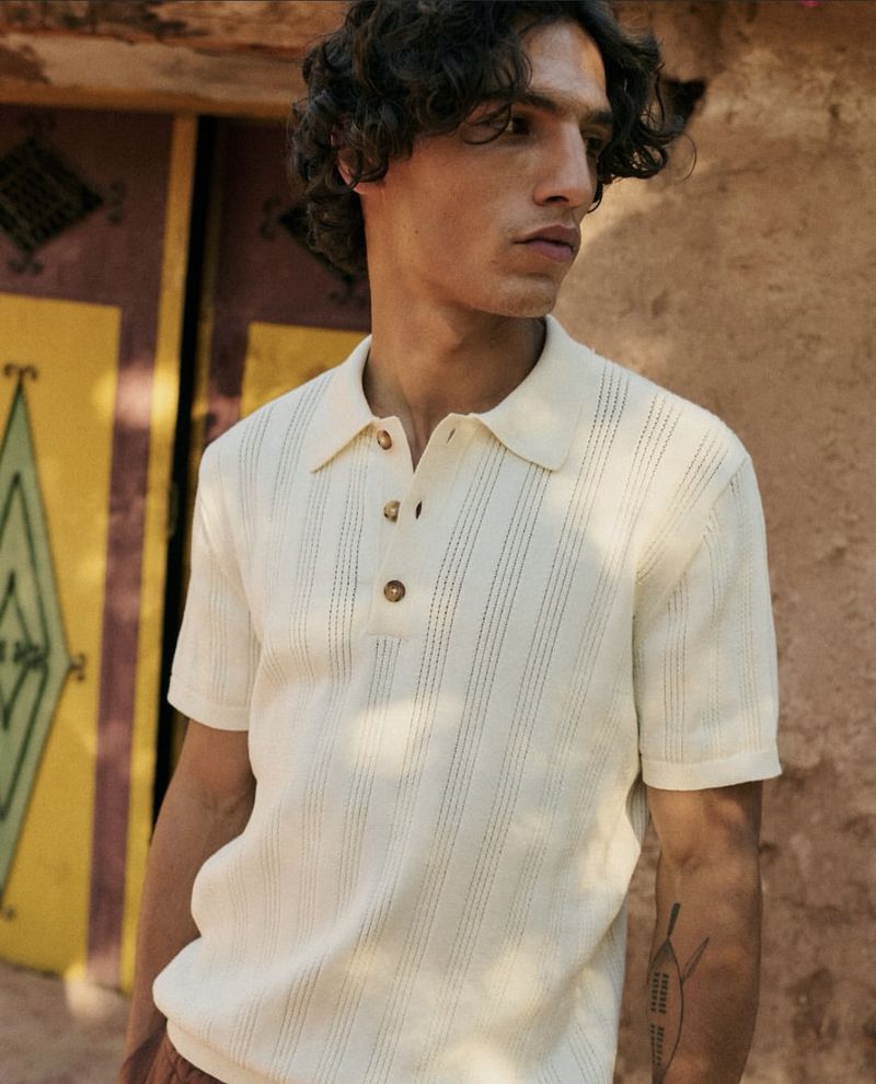 A man in a polo shirt stands outdoors against a painted door. He has curly brown hair, fair skin, and a tattoo on his forearm.