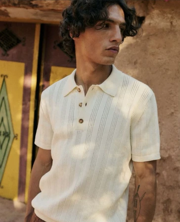 A man in a polo shirt stands outdoors against a painted door. He has curly brown hair, fair skin, and a tattoo on his forearm.