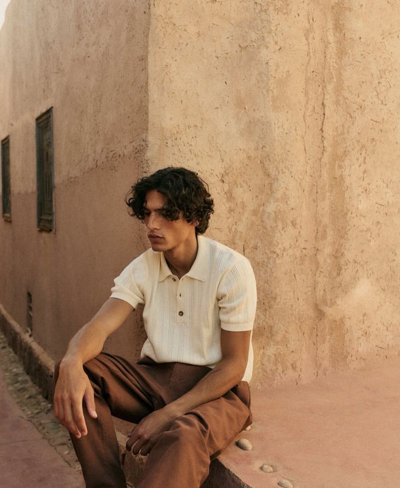 A young man with curly hair poses against a textured, adobe wall in a relaxed outdoor setting, wearing casual attire.
