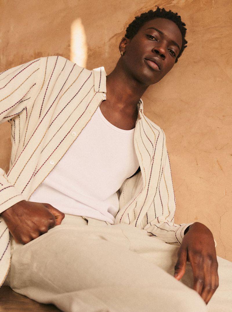 Dark-skinned male model poses in an open striped shirt and white undershirt, seated against a textured light-brown backdrop with natural lighting.