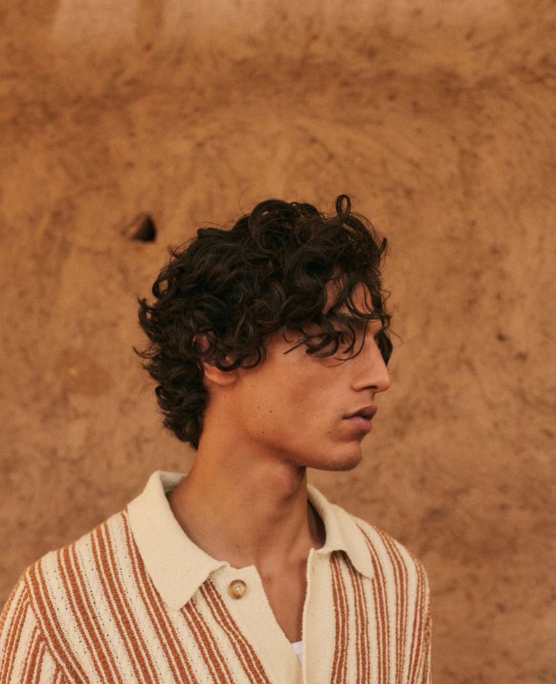 A young adult male model poses in a striped shirt in front of a plain tan wall, showcasing neutral colors and fashion details.