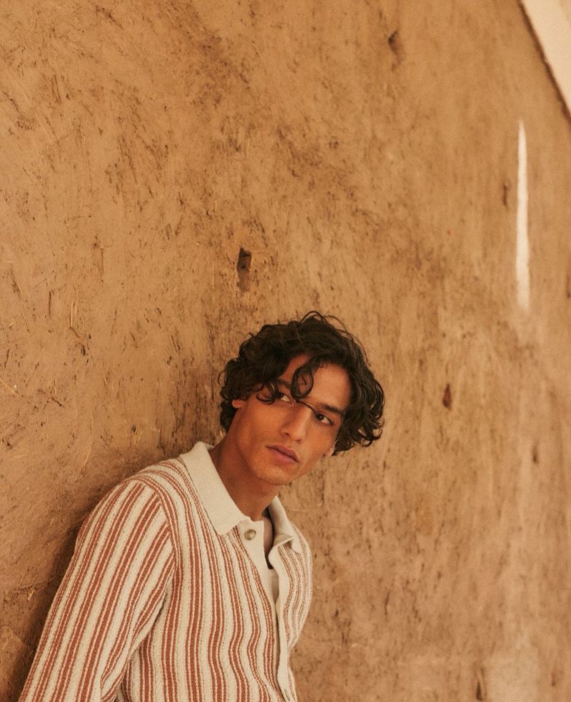 Portrait of a dark-haired man leaning against a textured wall, wearing a striped shirt in warm, neutral tones in a neutral setting.