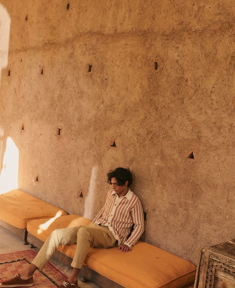 Person sitting on an orange bench inside a room with a brown wall. The person is wearing a striped shirt and light pants.