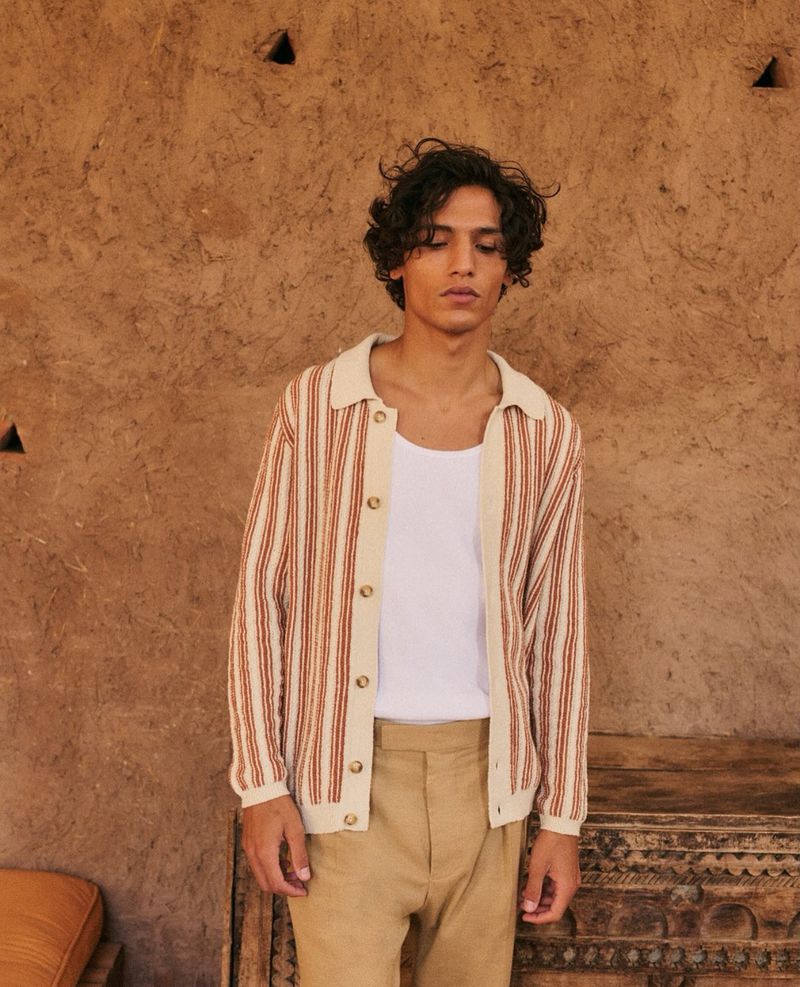 Adult male in a striped collared shirt and slacks stands indoors against a textured tan wall next to a carved wooden object.