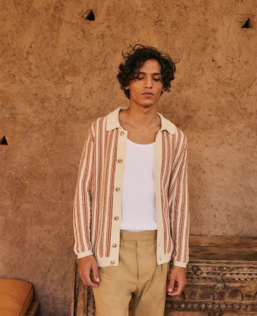 Adult male in a striped collared shirt and slacks stands indoors against a textured tan wall next to a carved wooden object.