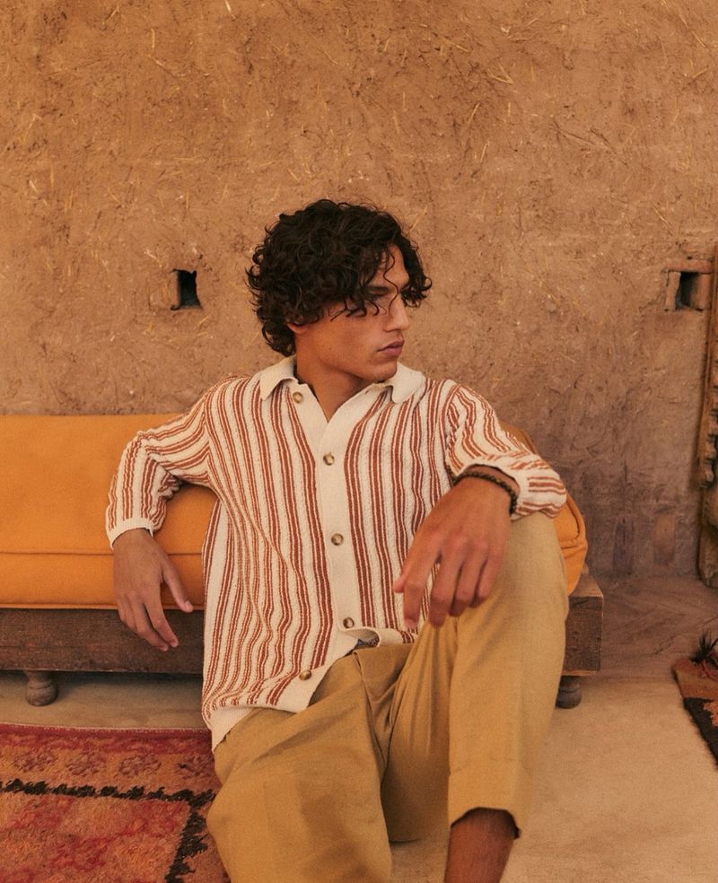 Young man with curly hair in striped shirt sitting against textured wall on orange cushioned bench, wearing khaki pants indoors.