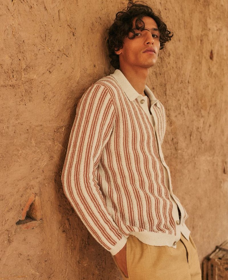 Portrait of a young adult man with curly hair wearing a striped knit shirt, leaning against a textured tan wall indoors, with natural light.