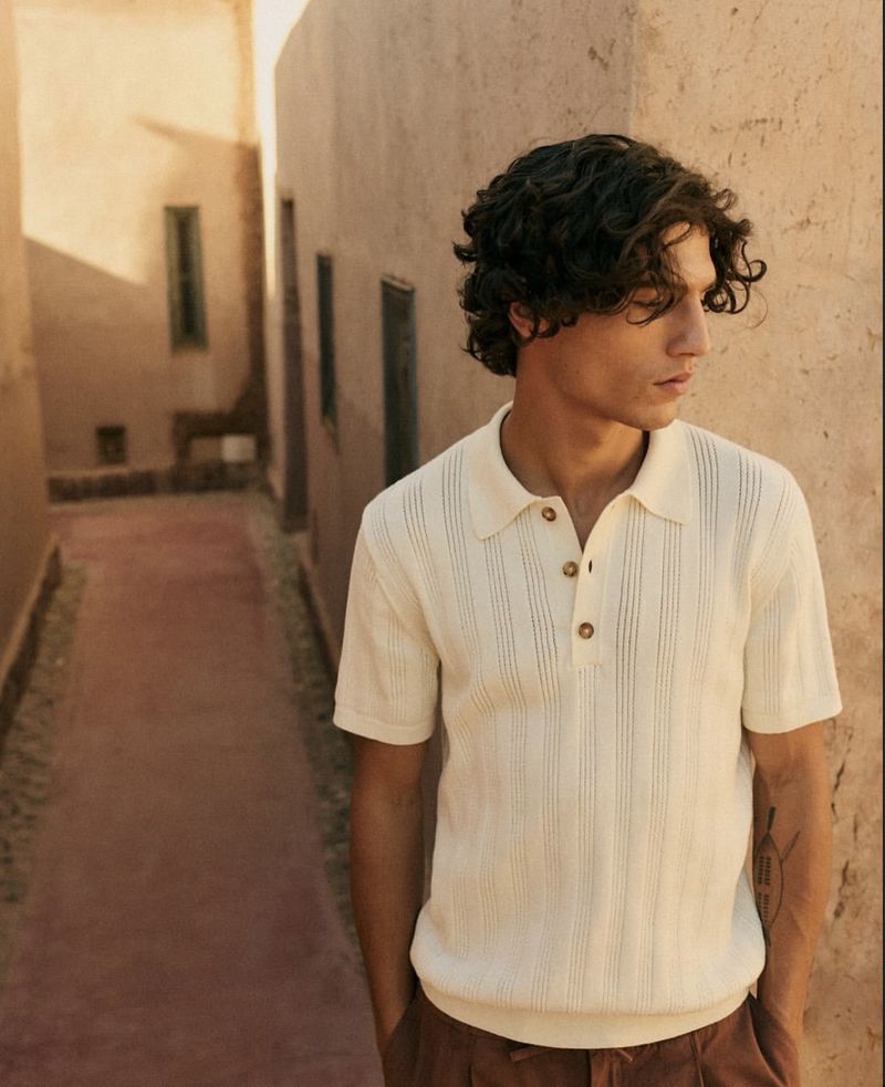 Young man with dark wavy hair in a cream-colored knit polo shirt poses in a tan alleyway with soft light and brown trousers.