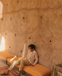 Model seated on a yellow cushioned bench against a textured earthen wall for Octobre Editions