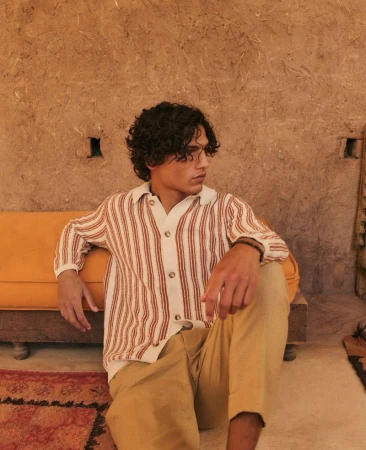 Man in a cream and brown striped knit buttoned polo shirt and camel trousers sitting against an ochre bench and textured wall.