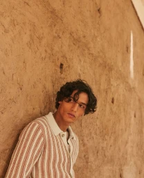 Model Morgan Sahadow wearing a striped knit polo shirt while leaning against a textured earth wall in warm natural light.